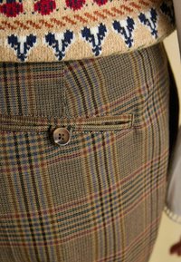 Checked trousers in brown, yellow, and green hues. Features a button closure with logo, side pocket, and woven textured fabric.