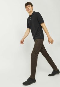 Polo shirt nero a maniche corte con tre bottoni, abbinato a pantaloni marroni slim-fit e scarpe nere stringate, che cammina su uno sfondo chiaro.