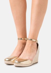 Nohy obuté do zlatých kovových wedge espadril s kotníkovými popruhy na jednoduchém světlém pozadí.