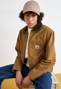 Chaqueta de lona marrón con un collar de pana, cremallera amarilla y un bolsillo en el pecho que presenta un logo. Una gorra beige clara completa el atuendo.