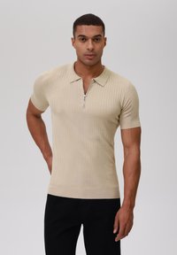 Beige geripptes Poloshirt mit kurzen Ärmeln, ausgestattet mit einem Reißverschluss auf der Vorderseite und tailliertem Schnitt. Getragen mit schwarzen Hosen, glatte Textur.