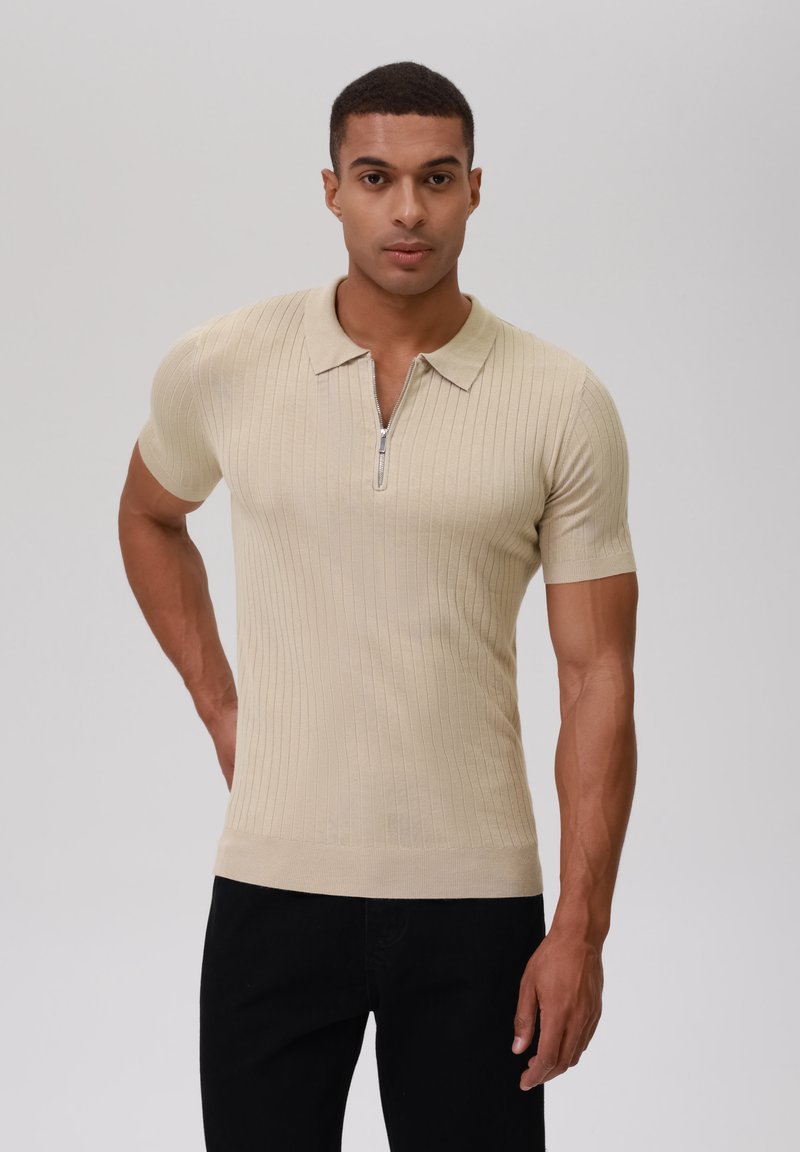 Beige geripptes Poloshirt mit kurzen Ärmeln, ausgestattet mit einem Reißverschluss auf der Vorderseite und tailliertem Schnitt. Getragen mit schwarzen Hosen, glatte Textur.