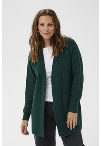 Grön stickad cardigan med långa ärmar, öppen front och en strukturerad yta. Tillsammans med en vit topp och mörka jeans.