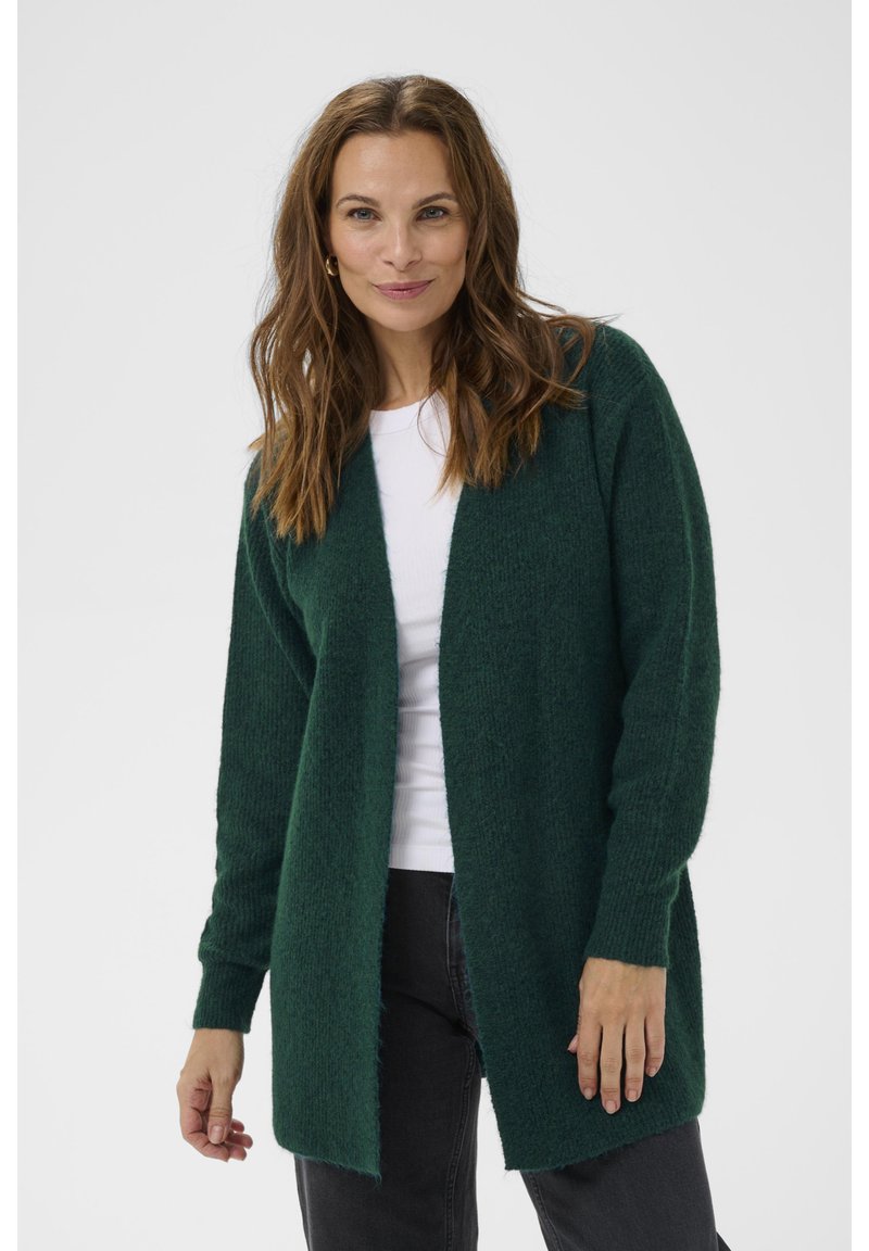 Grön stickad cardigan med långa ärmar, öppen front och en strukturerad yta. Tillsammans med en vit topp och mörka jeans.