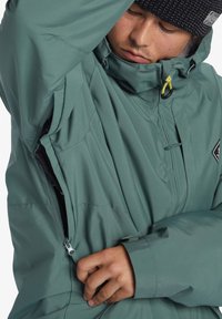 Svetlozelená vodoodolná bunda s vysokým golierom, zipsovými ventilmi a textúrovanou čiernou čiapkou. Má priliehavý strih a nastaviteľný lem.