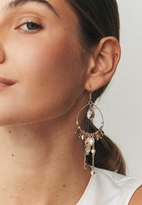 Gold hoop earring with a teardrop-shaped crystal, pearl accents, and layered chains featuring leaf charms, displaying intricate detailing.