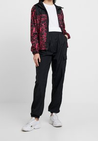 Czarna i różowa kurtka wiatrówka w wzory, lekki materiał, zapięcie na zamek, zestawiona z czarnymi spodniami typu cargo i białymi trampkami.