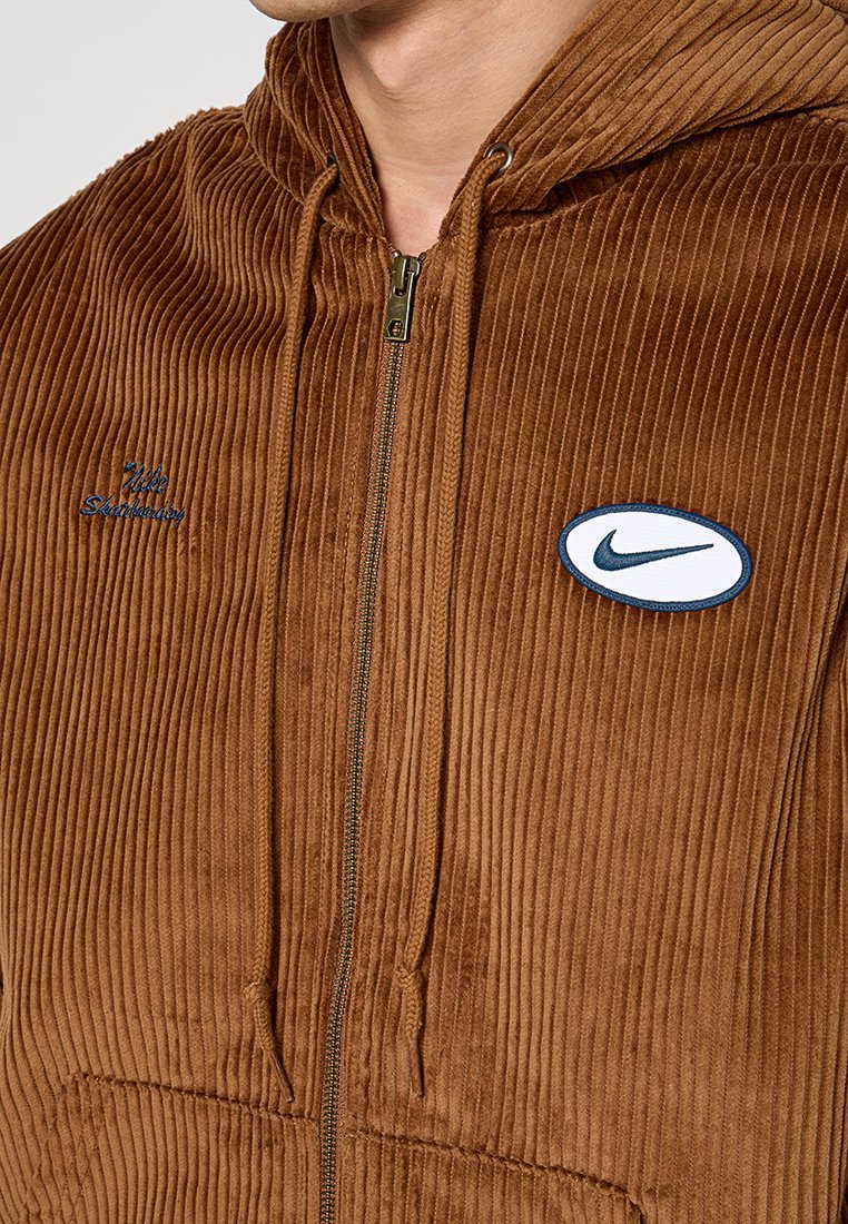 Sudadera con cremallera de pana marrón, con un bolsillo frontal, capucha con cordón, parche del logo de Nike y textura acanalada. Acentos bordados en azul.