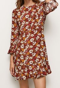 Kvinna i en långärmad klänning ovanför knäet med ett blommigt mönster i rött, gult och vitt samt volanger vid fållen och ärmsluten.