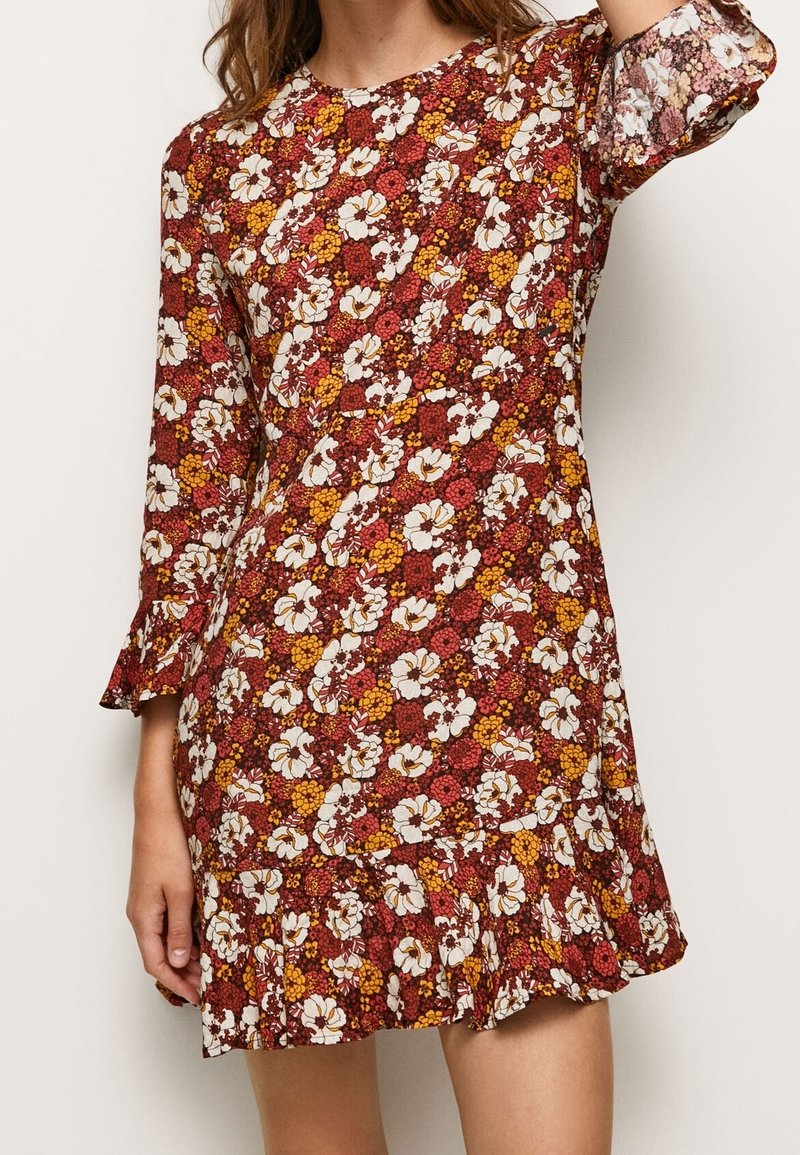 Kvinna i en långärmad klänning ovanför knäet med ett blommigt mönster i rött, gult och vitt samt volanger vid fållen och ärmsluten.