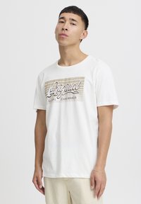 Bílý bavlněný tričko s grafickým designem, který na přední straně zobrazuje text "Original Global Jeansmaker" v taupe a černé barvě. Krátké rukávy, kulatý výstřih.
