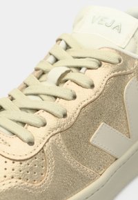 Gros plan d'une basket Veja beige avec des panneaux texturés, des lacets crème et un logo sur le côté et la languette.