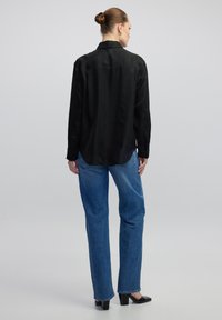 Schwarze Bluse mit langen Ärmeln und glatter Textur, mit einem Rückenschlitz. Kombiniert mit blauen Jeans im geraden Schnitt und schwarzen Absatzschuhen.