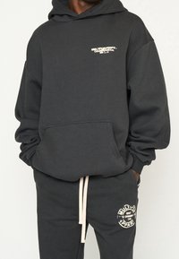 Sweat-shirt à capuche noire en matériau doux, avec une poche avant et un petit logo sur la poitrine. Assorti à un pantalon de jogging avec un détail de patch.
