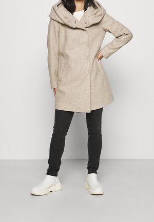 Femme portant un manteau à capuche beige, un pantalon noir slim et des bottines blanches, debout avec une main sur la hanche sur un fond uni.
