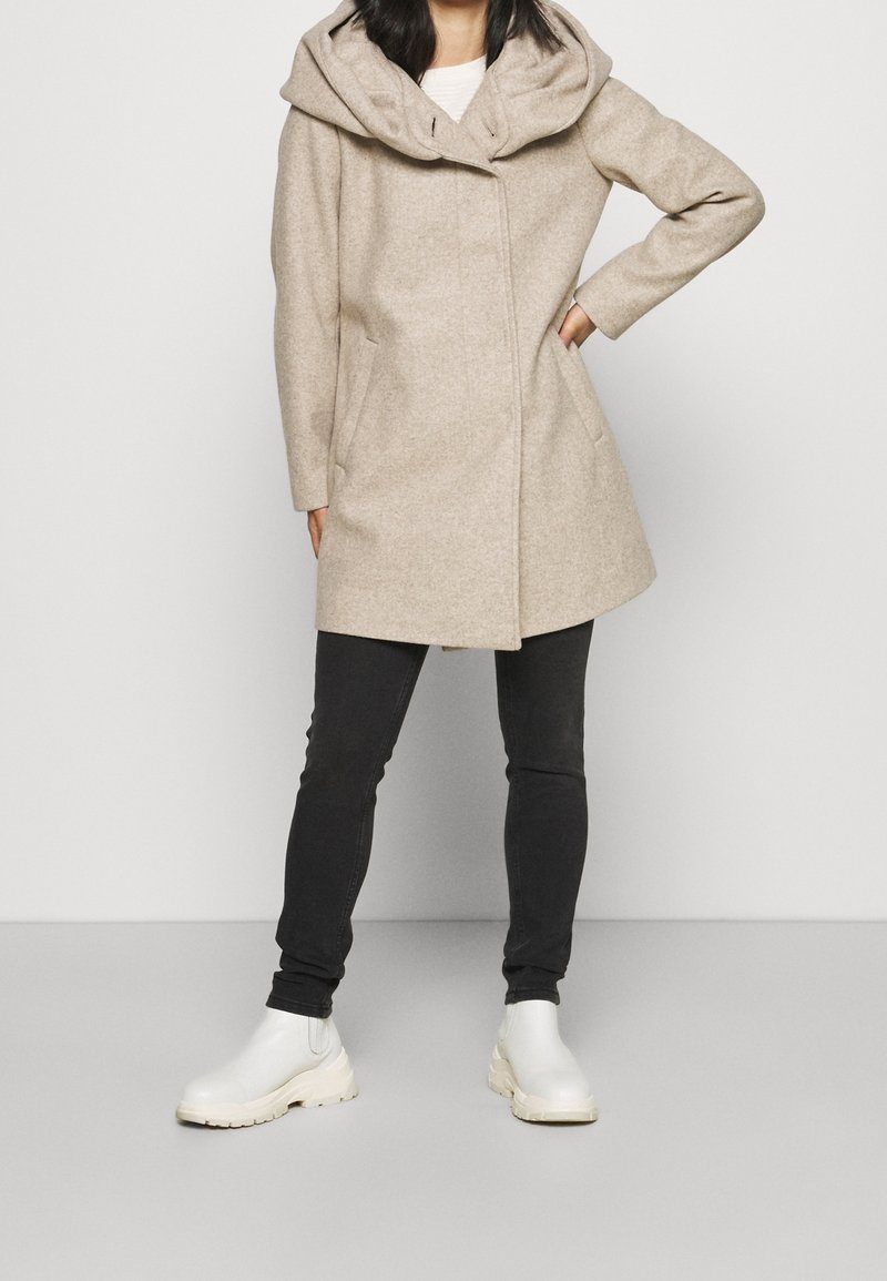 Femme portant un manteau à capuche beige, un pantalon noir slim et des bottines blanches, debout avec une main sur la hanche sur un fond uni.