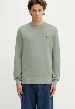 Giovane con capelli corti e ricci che indossa un maglione a maniche lunghe verde chiaro e jeans blu, in piedi contro uno sfondo neutro.