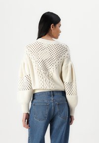 Pull en tricot couleur crème avec des motifs de câbles texturés et des détails en tricot ajouré sur les épaules et les manches, coupe courte et ourlet côtelé.