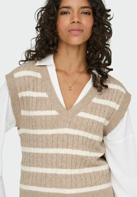 V-kauluksellinen, hihaton beige neulepaita, jossa on valkoisia raitoja; sisältää palmikkoneulekuvion ja ribbauseristet. Päällä valkoisen kauluspaitan päällä.