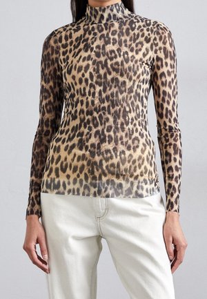 Vrouw draagt een nauwsluitende lange mouwen top met luipaardprint en een hoge hals, gecombineerd met een witte broek, tegen een effen grijze achtergrond.