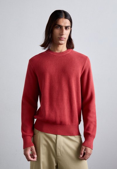 Rød strikket sweater med rund hals og ribbede manchetter og kant. Båret over beige bukser med en struktureret, let design.