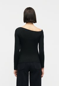 Γυναίκα με κοντά, ίσια μαλλιά φοράει μαύρο τοπ με μακριά μανίκια, χωρίς ώμους και μαύρο παντελόνι, στέκεται με την πλάτη γυρισμένη σε απλό φόντο.