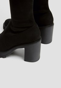 Botas de ante negras hasta la mitad de la pantorrilla con tacones gruesos y suelas de goma con relieve sobre un fondo gris claro.