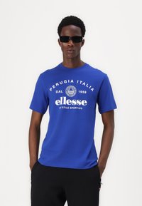 T-shirt de algodão azul com um logótipo e texto impressos em branco. O modelo usa óculos de sol pretos. Decote redondo simples e mangas curtas.