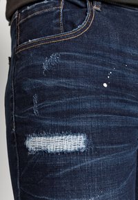 Jeans de mezclilla oscura con parches desgastados, incluyendo bordes deshilachados y salpicaduras de pintura blanca, con costuras amarillas a lo largo de las uniones.