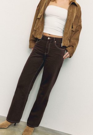 Frau trägt eine braune Wildlederjacke, ein weißes trägerloses Oberteil, dunkelbraune Jeans und beige Stiefel mit Absatz, steht vor einer weißen Wand.