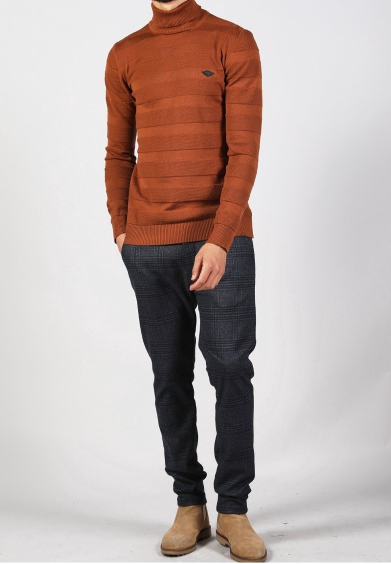 Homme portant un pull à col roulé couleur rouille, un pantalon à carreaux foncés et des bottes en daim beige, debout les mains détendues.