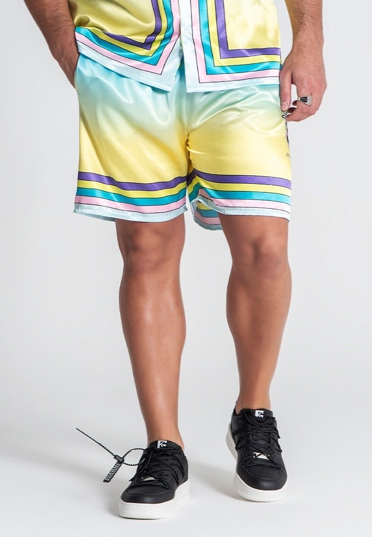 Parte inferiore del corpo di una persona che indossa pantaloncini a righe lucide di colori pastello e una maglietta abbinata, con scarpe da ginnastica nere e un anello sulla mano destra.