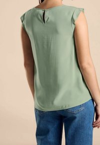 Blouse verte sans manches avec une ouverture en forme de goutte d'eau dans le dos, fabriquée en tissu doux. Associée à un jean en denim bleu, vue de dos.