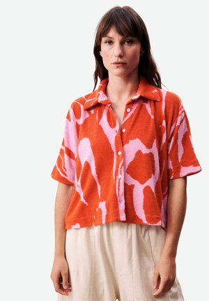 Vrouw draagt een oranje-roze abstract patroon korte mouwen overhemd met knopen en beige hoog getailleerde broek tegen een witte achtergrond.