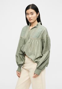 Γυναίκα με μακριά σκούρα μαλλιά φοράει ανοιχτό πράσινο κεντητό πουκάμισο με πιετέ μανίκια και κρεμ παντελόνι ψηλόμεσο, στέκεται μπροστά σε λευκό φόντο.