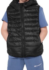 Černá péřová vesta s kapucí, se zipem vpředu a žebrovanou strukturou. Nasažená přes světle modré tričko a doplněná o modré šortky stejné barvy.