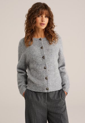 Cardigan grigio in lana con collo rotondo, sei bottoni scuri e polsini a coste. Indossato sopra pantaloni grigi a righe, con una finitura testurizzata.