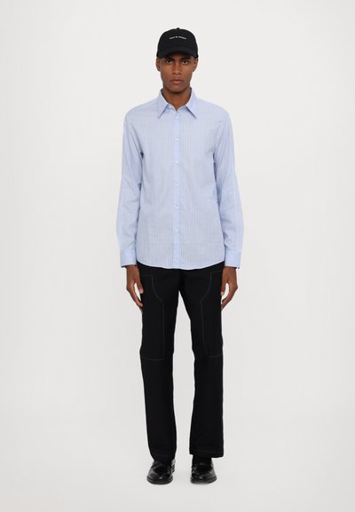 Hellblau gestreiftes Hemd mit Knöpfen, schwarze Hose mit Nähdetails und schwarze Kappe mit weißem Text. Minimalistisches Design, figurbetonter Schnitt.