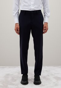 Man in zwart slim-fit broek, wit overhemd met knopen, en zwart instap sneakers op een lichtgrijs tapijt.