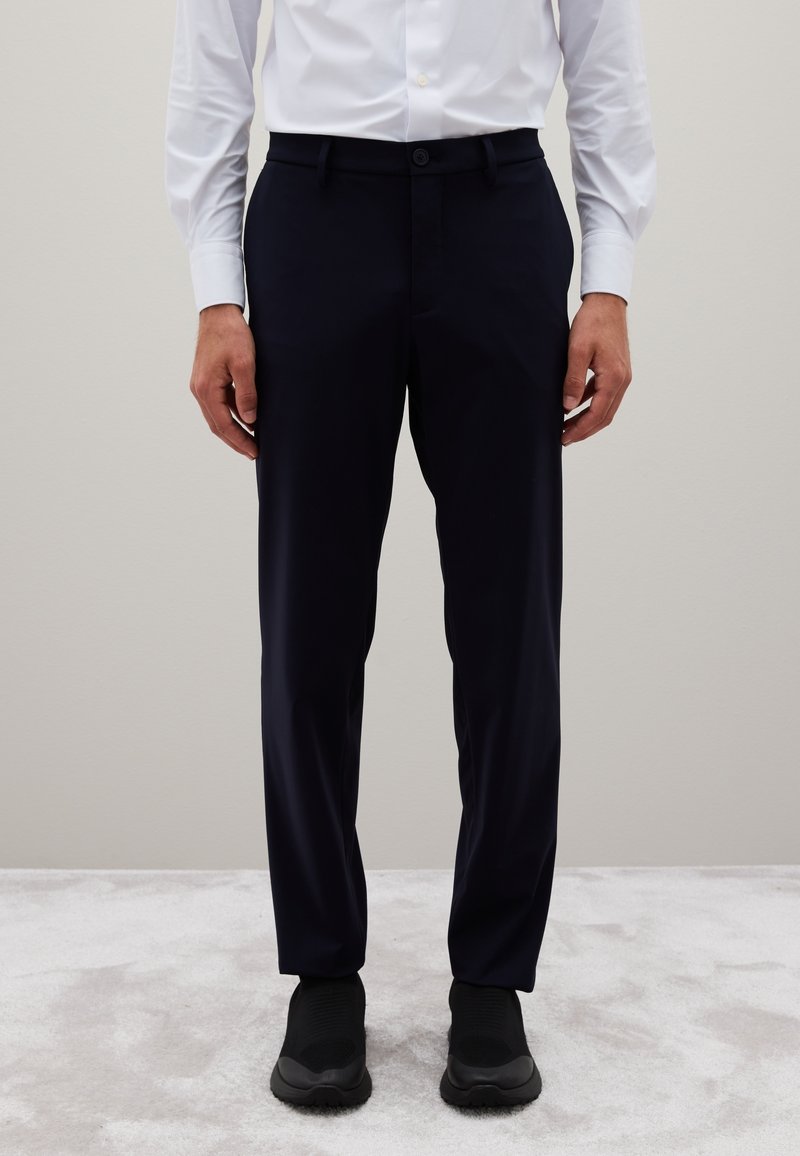 Man in zwart slim-fit broek, wit overhemd met knopen, en zwart instap sneakers op een lichtgrijs tapijt.