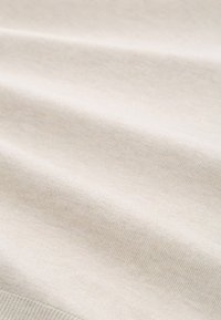 Beige katoenen stof met een zachte textuur, voorzien van een fijne ribbelpatroon. Het materiaal valt soepel en toont subtiele plooien.