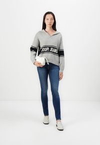 Pull en tricot gris avec capuche, présentant une rayure logo "JOOP! JEANS" en noir et blanc. Porté avec un jean bleu et des chaussures blanches à enfiler.