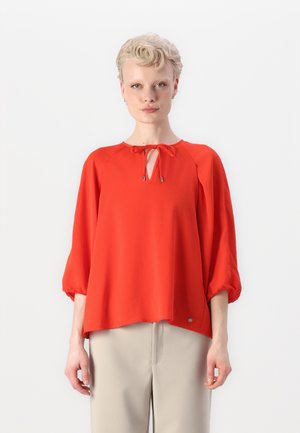 Rød bluse med rund halslinning, knyte detalj og trekvartlange ermer. Jevnt stoff og løs passform; liten logo aksent ved sømmen.