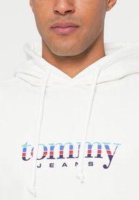 Λευκή hoodie με πολύχρωμο λογότυπο "tommy jeans". Το υλικό φαίνεται μαλακό με κορδόνια και σχέδιο τσέπης καγκουρό.