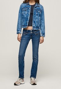 Persona vestida con chaqueta de denim azul, top de corte corto oscuro, jeans azules con doble botón y zapatillas beige con acentos grises y rosas.