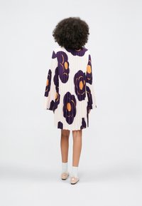 Žena s kučeravými vlasmi má na sebe plisované biele šaty s veľkými fialovými a oranžovými kvetinovými vzormi, biele ponožky a ružové topánky, stojí pred jednoduchým pozadím.