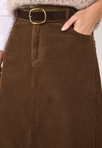 Gonna in velluto a coste marrone con costine verticali, tasche frontali e cintura marrone scuro con fibbia rotonda dorata e cuciture decorative.