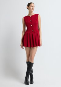 Mujer vestida con un mini vestido rojo sin mangas con botones dorados y falda plisada, combinado con botas de cuero negras hasta la rodilla, apoyada contra una pared blanca.