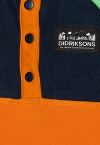 Giacca in pile con parte superiore blu navy, sezione inferiore arancione e patta dei bottoni arancione. Presenta un'etichetta nera con la scritta "DIDRIKSONS" e un grafico.