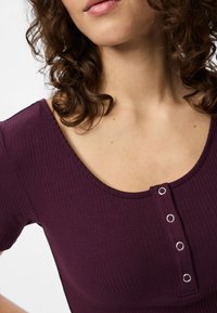 Artimas plano nuotrauka, kurioje matoma moteris su garbanotais plaukais, vilkinti tamsiai violetinį, ripsinį megztinį su apvalia iškirpte ir keturiais metaliniais mygtukais priekyje.